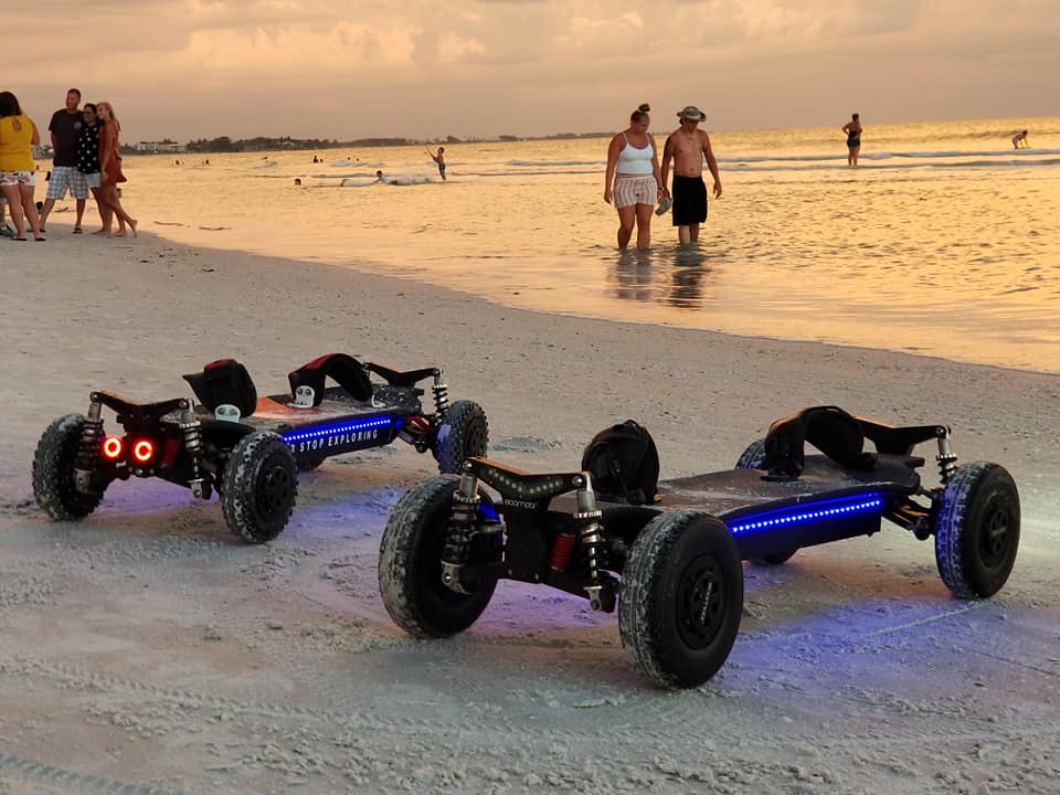 Two electric mountainboards with blue LED underglow and red taillights parked on a sandy beach at sunset, with people in the background.