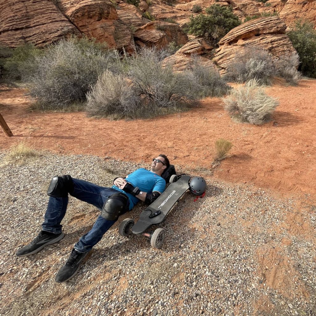 A person relaxing on a DIY electric skateboard in a desert landscape with red rock formations and sparse vegetation.