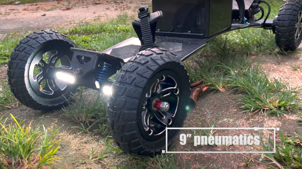 Electric mountainboard with 9-inch pneumatic tires, showing close-up of wheel and lights on a dirt path.