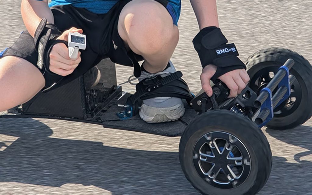 Person crouching on an electric skateboard, wearing protective gear.